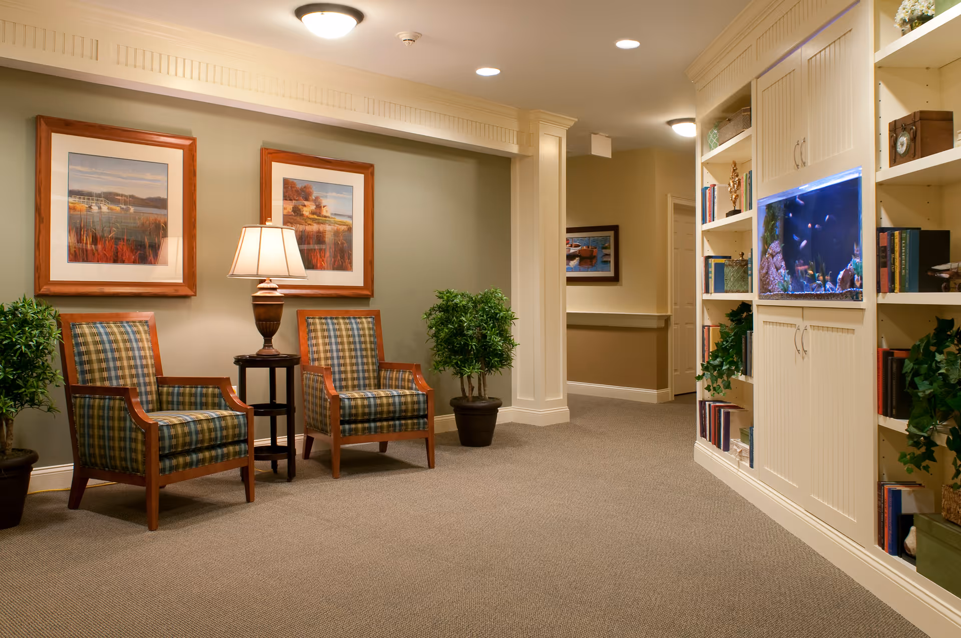 A cozy interior common area with two plaid upholstered wooden armchairs separated by a small table with a lamp. Two framed landscape paintings hang on the wall behind the chairs. There are potted plants on either side of the chairs. To the right, built-in white shelves hold books, decorative items, and a large aquarium with colorful fish. The space is warmly lit with ceiling lights and has beige carpeting.