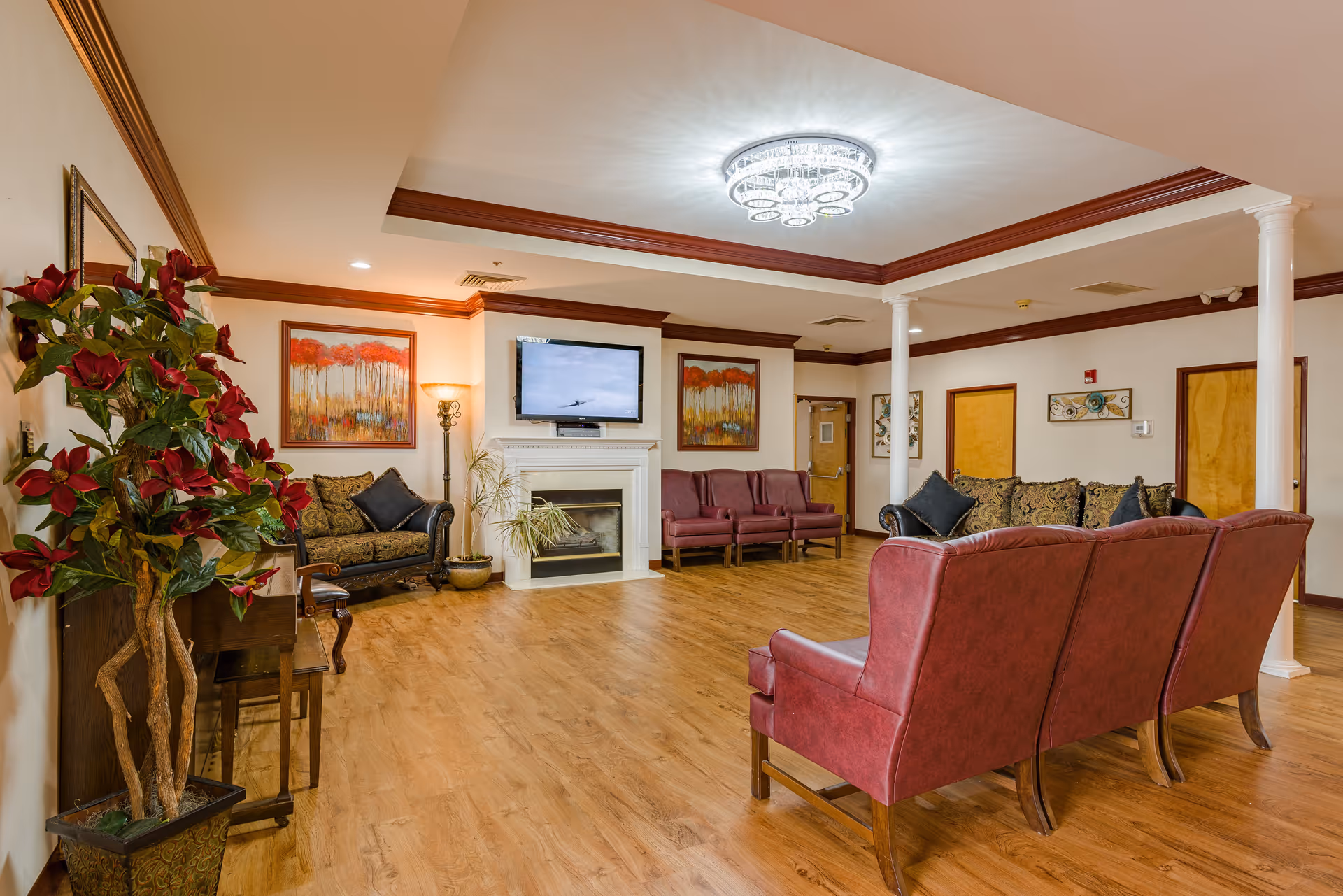 A spacious and well-lit living room in Brentwood Retirement Community featuring wooden flooring, a white fireplace with a mounted flat-screen TV above it, multiple seating options including red armchairs and a patterned sofa, decorative paintings on the walls, a tall floor lamp, and a large potted plant with red flowers near a wooden piano.