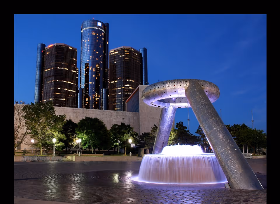 A illuminated circular fountain in a public plaza at night with tall glass skyscrapers in the background.