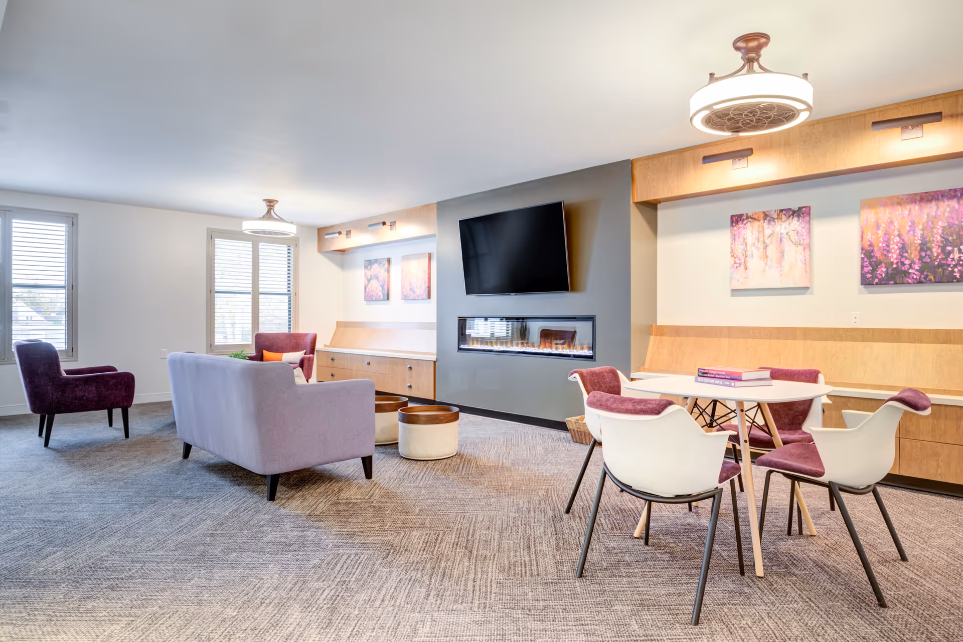 Bright communal lounge with sofas and chairs, a wall-mounted TV above a linear fireplace, and a small dining table with chairs.