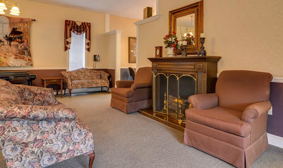 A cozy living room area in Rock Point Nursing Center featuring two upholstered armchairs and two floral-patterned sofas arranged around a wooden fireplace with a brass screen. The fireplace mantel is decorated with a clock, candles, and a floral arrangement, and a large mirror hangs above it. A piano and a small side table are visible in the background near a window with a valance.