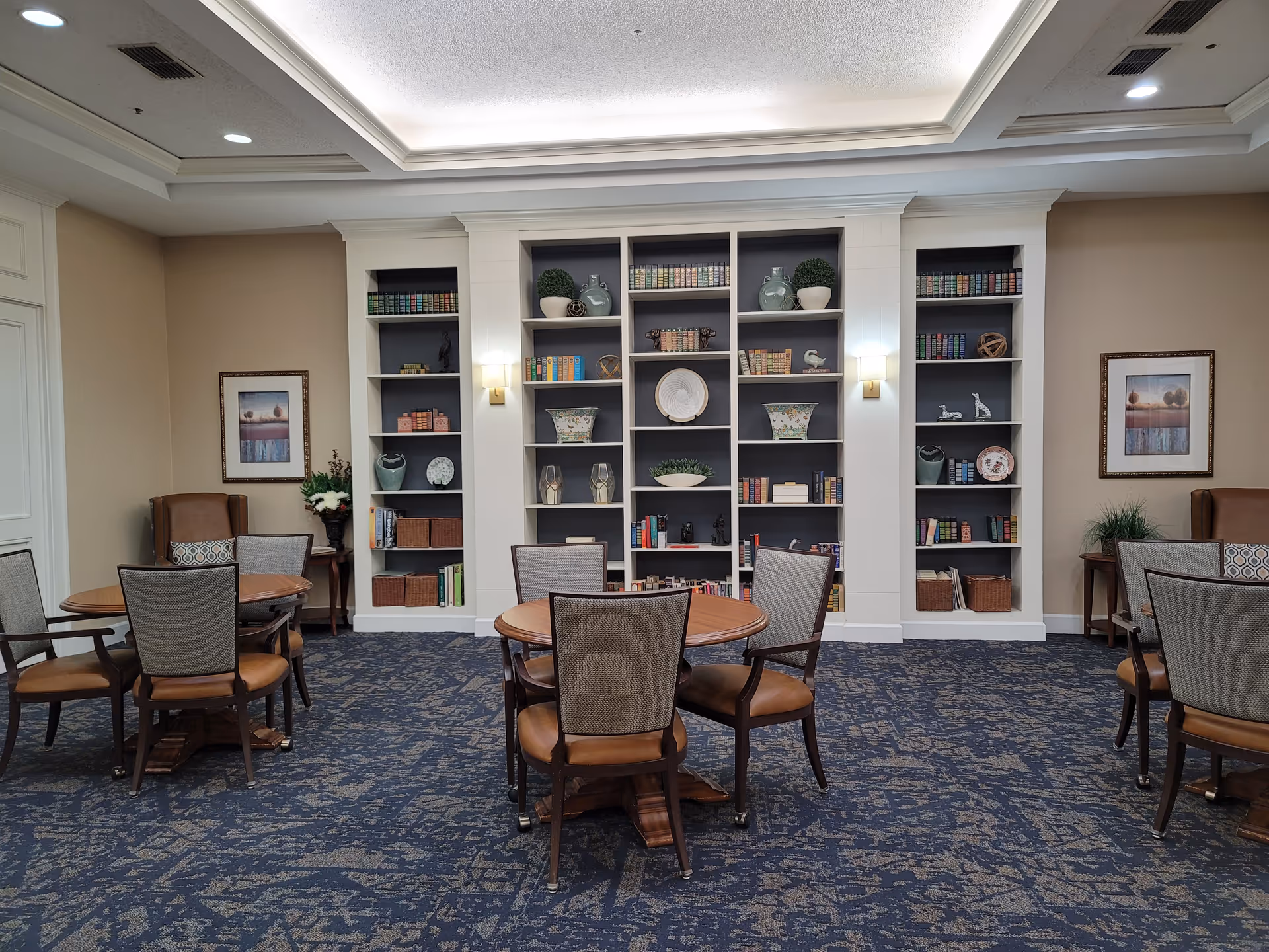 A cozy common area with round wooden tables and cushioned chairs arranged on a patterned blue carpet. The back wall features built-in shelves filled with decorative items, books, and plants. Two framed pictures hang on the beige walls, and there are two armchairs with side tables and plants in the corners.