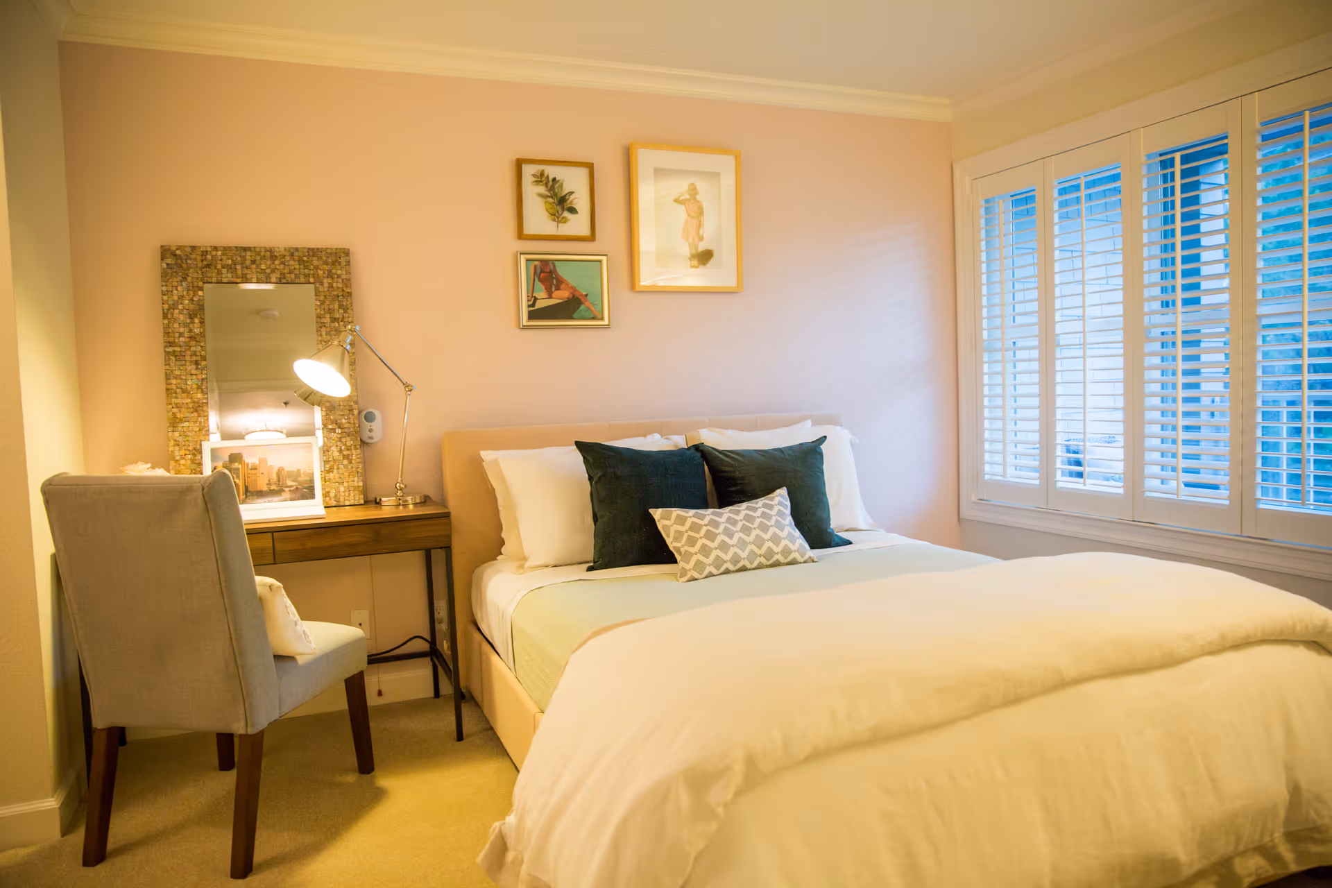 A cozy bedroom with a neatly made bed featuring white and green pillows and a white comforter. There is a wooden desk with a decorative mirror, a lamp, and framed pictures on the wall above the bed. A cushioned chair is placed next to the desk, and a window with white shutters allows natural light into the room.