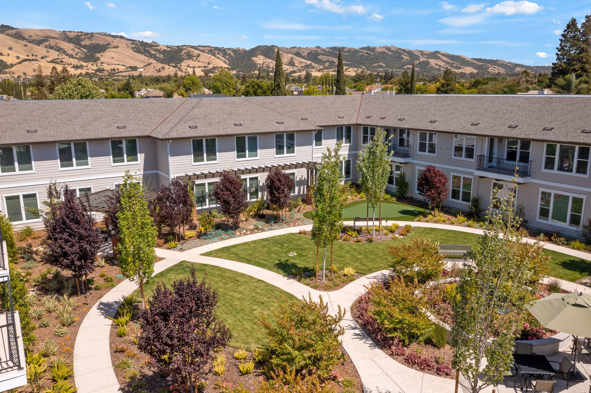 A landscaped courtyard of a senior living facility with walking paths, green lawns, various trees and shrubs, outdoor seating with tables and umbrellas, surrounded by a two-story building with multiple windows and balconies. Hills and a blue sky with some clouds are visible in the background.