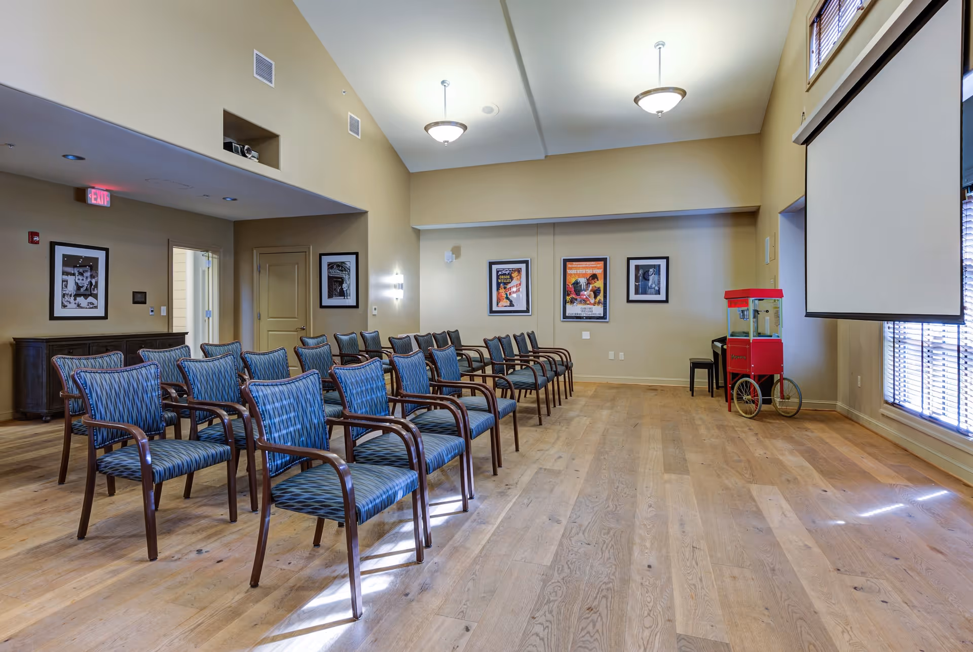 A room set up with rows of blue cushioned chairs with wooden armrests facing a large white projection screen. The room has wooden flooring, beige walls, framed pictures on the walls, and a red popcorn machine near the window with blinds.