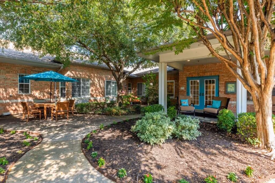 Outdoor courtyard area of Spring Creek Assisted Living featuring a paved walkway, wooden table with chairs and a blue umbrella, surrounded by trees and shrubs. There is a covered seating area with two cushioned chairs and a small table near blue double doors on a brick building.