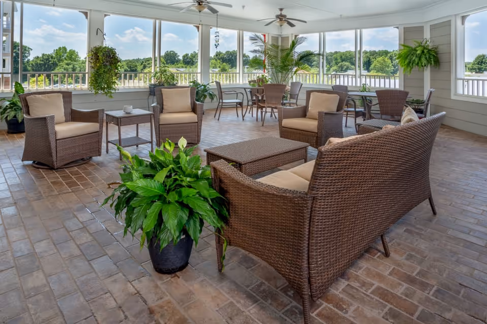 A bright and spacious enclosed patio area with wicker furniture including a sofa, armchairs, and tables. The floor is covered with brick tiles, and there are several green plants placed around the space. Large windows offer a view of trees and a blue sky outside. Ceiling fans are mounted on the white ceiling.