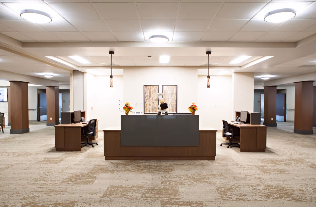A spacious reception area with a central desk featuring a dark front panel and two side desks with office chairs and computer monitors. The area is well-lit with ceiling lights and pendant lights above the central desk. There are two framed artworks on the wall behind the desk and flower arrangements on either side. The floor is carpeted with a patterned design, and the space extends into hallways with wooden columns and doors.