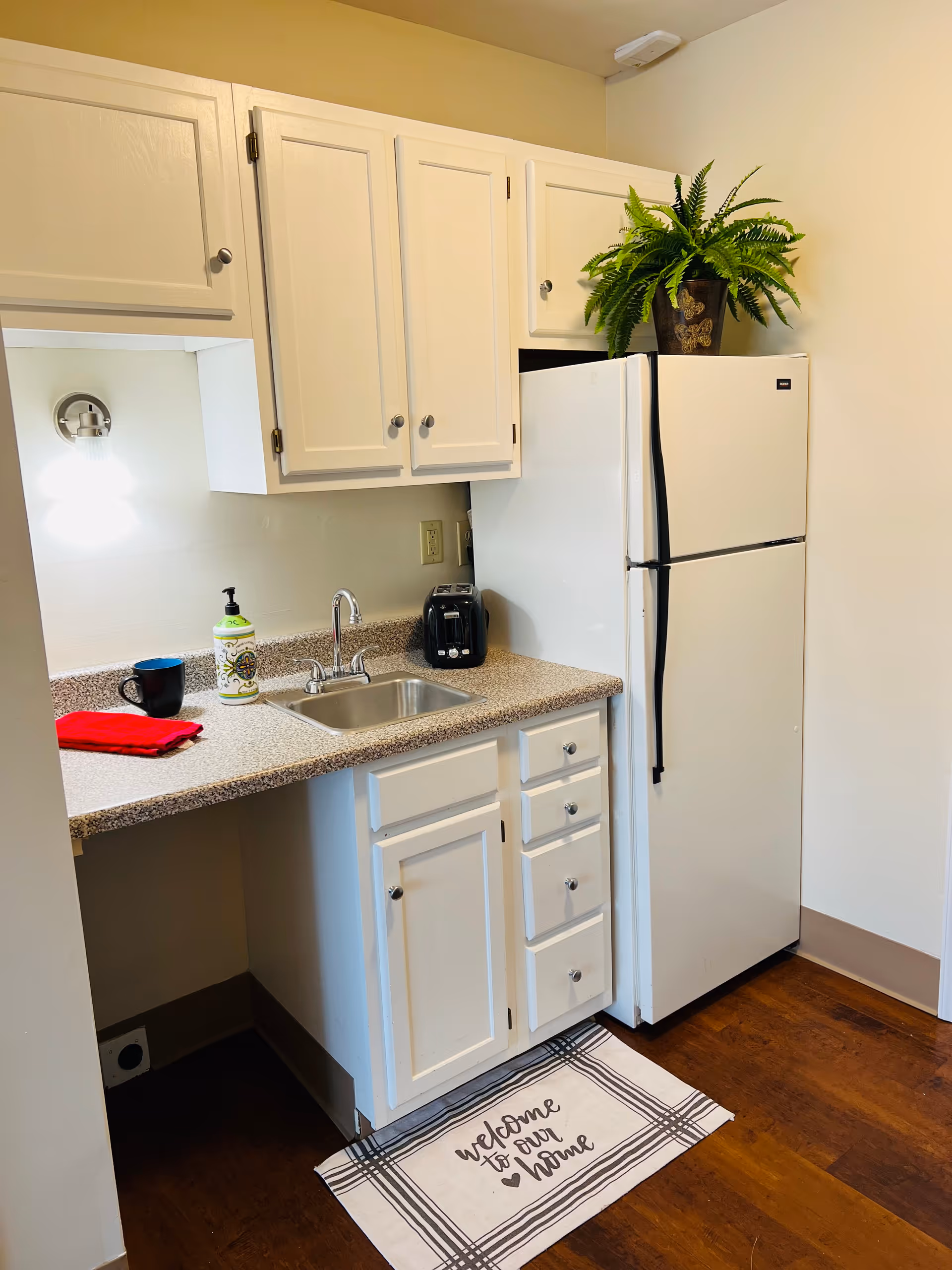 A small kitchen area with white cabinets, a countertop with a sink, a black toaster, a soap dispenser, a black mug, and a red cloth. There is a white refrigerator with a green potted plant on top. A light fixture is mounted on the wall above the countertop, and a floor mat with the text 'welcome to our home' is placed on the wooden floor.