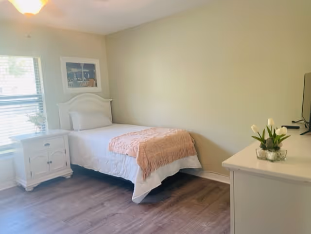 A simple, clean bedroom with a single bed made with white bedding and a pink throw blanket. There is a white nightstand with a small plant next to the bed, a window with blinds letting in natural light, a framed picture on the wall above the bed, and a white dresser with a small vase of flowers and a TV on top.