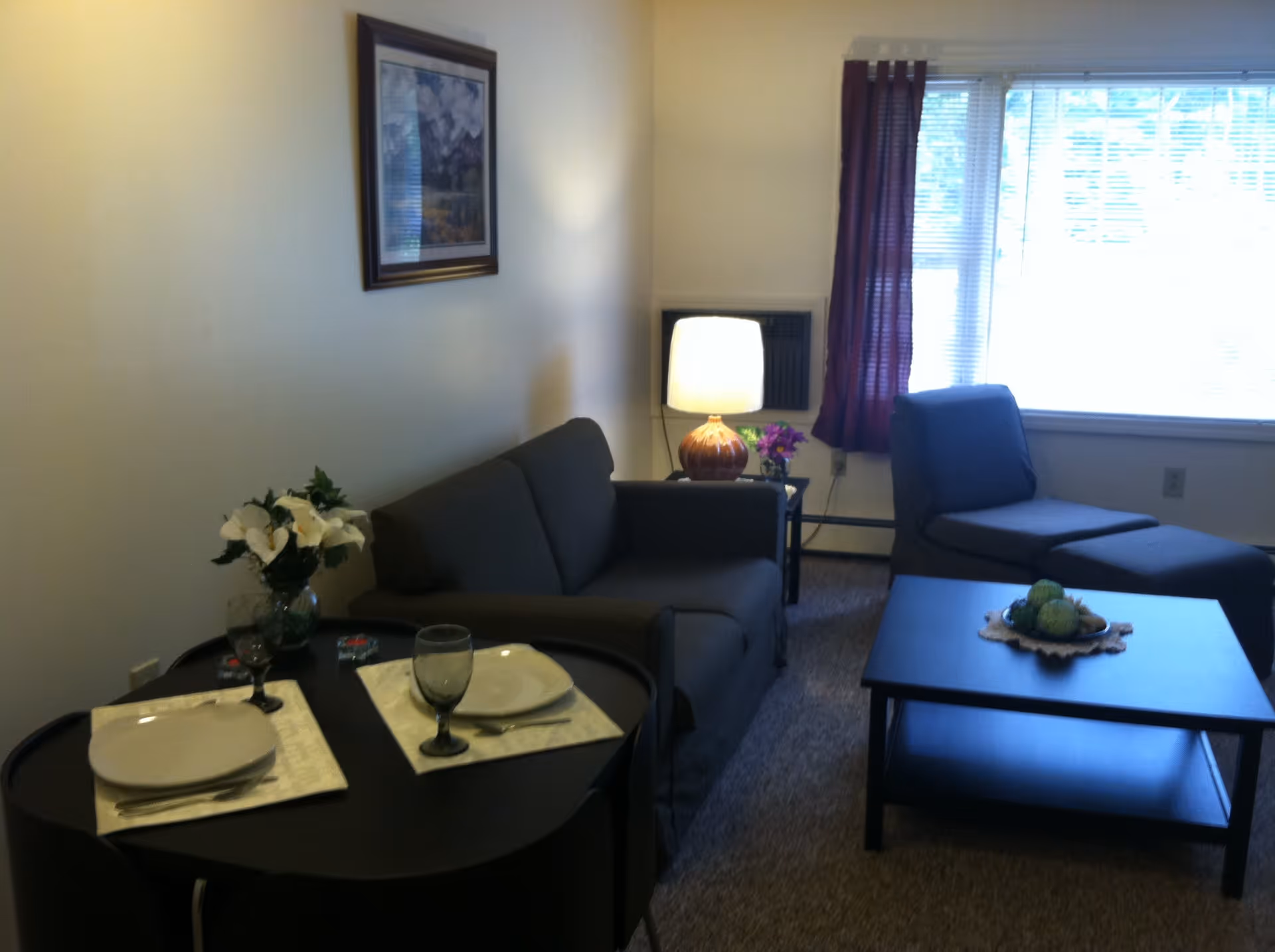 A small living room area with a dark gray sofa and matching chaise lounge near a window with blinds and purple curtains. A black coffee table with a decorative centerpiece is in front of the sofa. To the left, there is a small round dining table set for two with white plates, glasses, and a vase of white flowers. A framed picture hangs on the wall above the sofa, and a table lamp with a round base is on a side table next to the sofa.