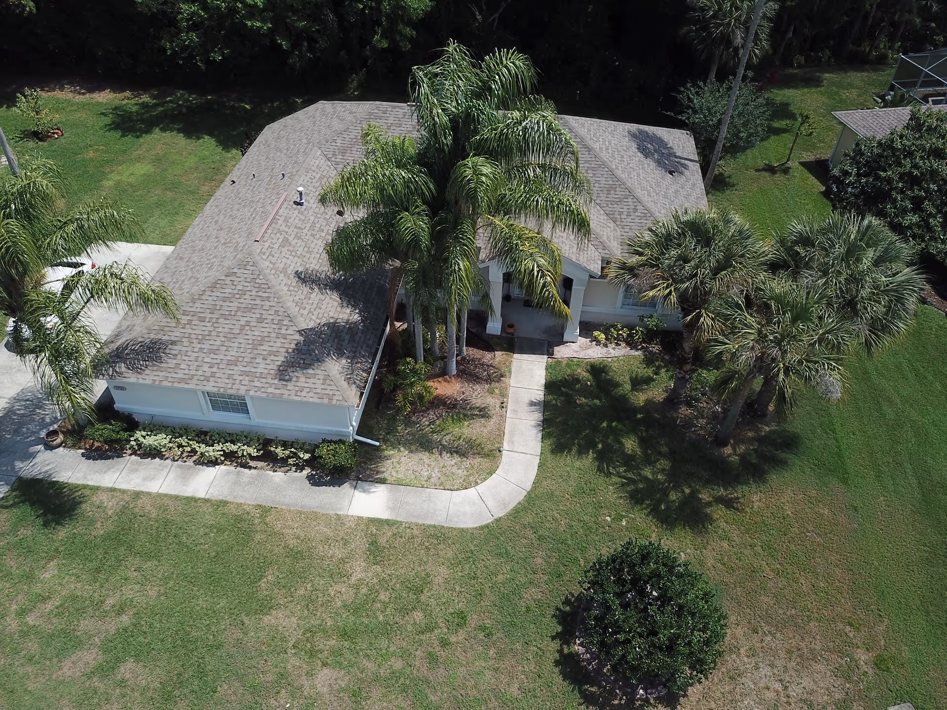 Aerial view of a single-story house with a shingled roof, palm trees and a curved walkway across a lawn.