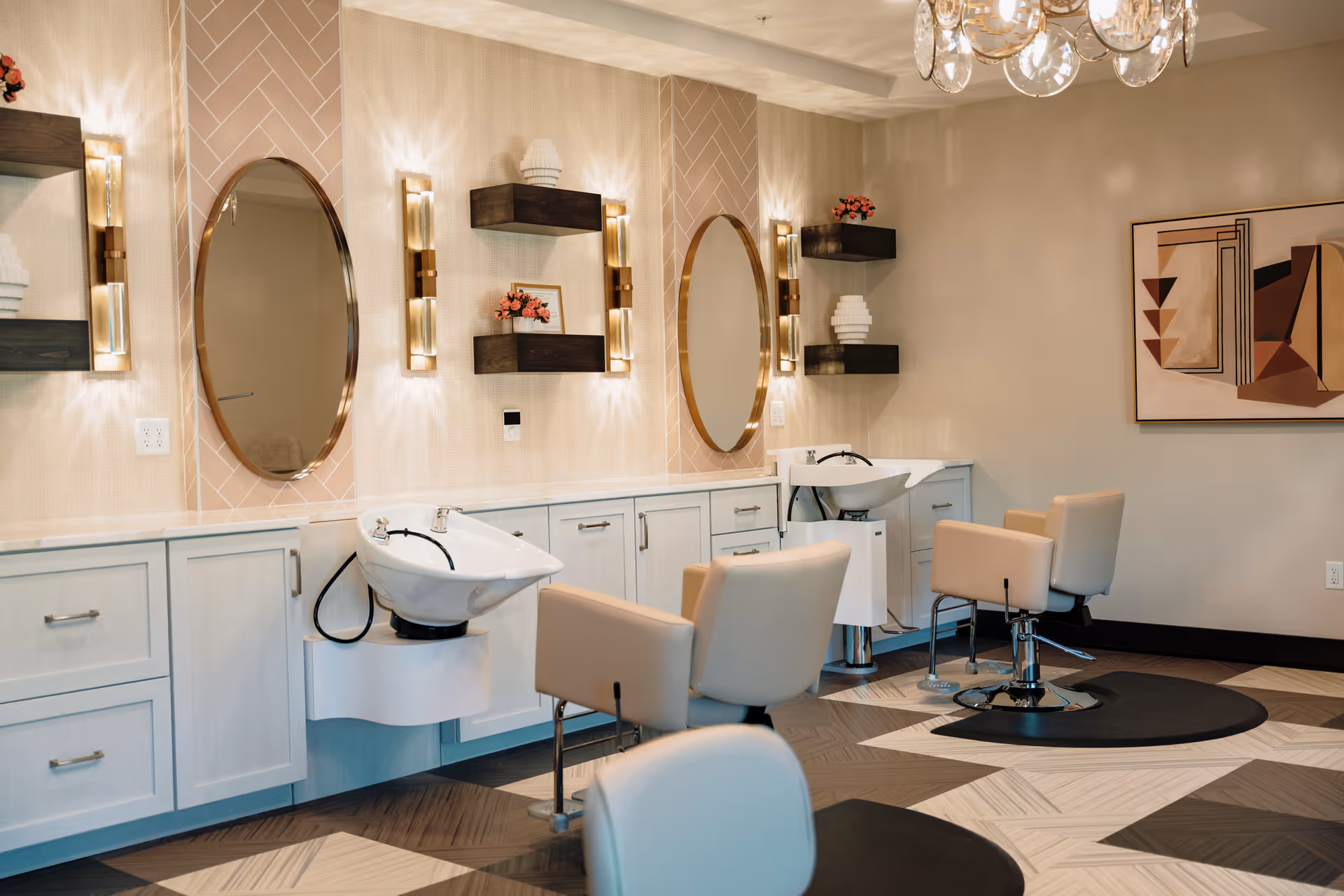 Interior of a modern hair salon area with two beige salon chairs in front of white wash basins. The wall features two oval mirrors with decorative wall sconces and floating shelves holding small plants and decor items. The floor has a geometric patterned design in neutral tones, and a contemporary chandelier hangs from the ceiling.