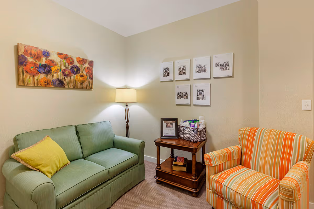 A cozy sitting area with a green couch featuring a yellow pillow, an orange and white striped armchair, a wooden side table with a basket of supplies and framed picture, a floor lamp, and colorful floral artwork on the wall.