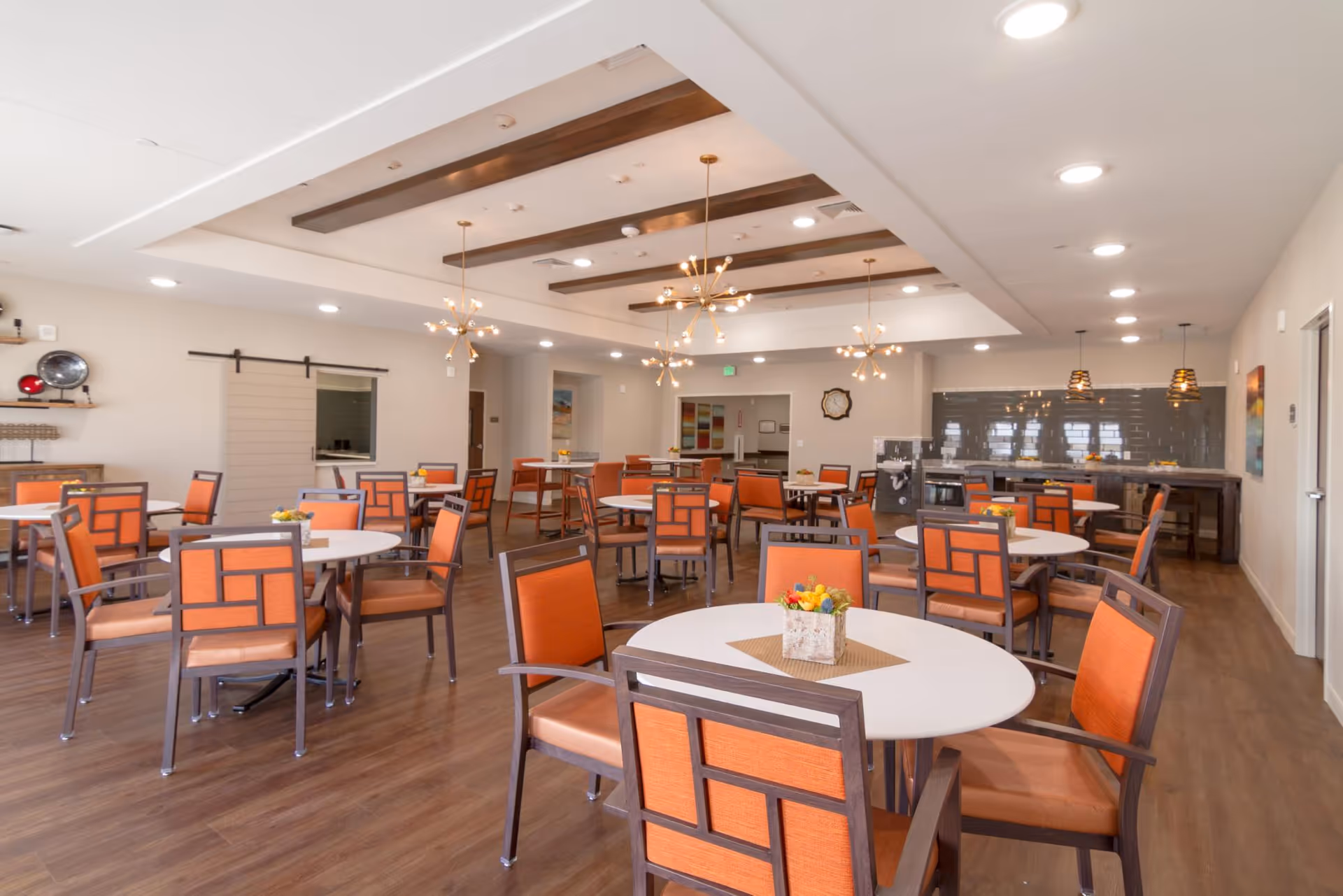 A spacious dining room with multiple round and square tables surrounded by orange cushioned chairs. The room features modern pendant lighting, wooden ceiling beams, and a serving counter with a dark tiled backsplash. The floor is wooden, and the walls are painted a light color, creating a warm and inviting atmosphere.