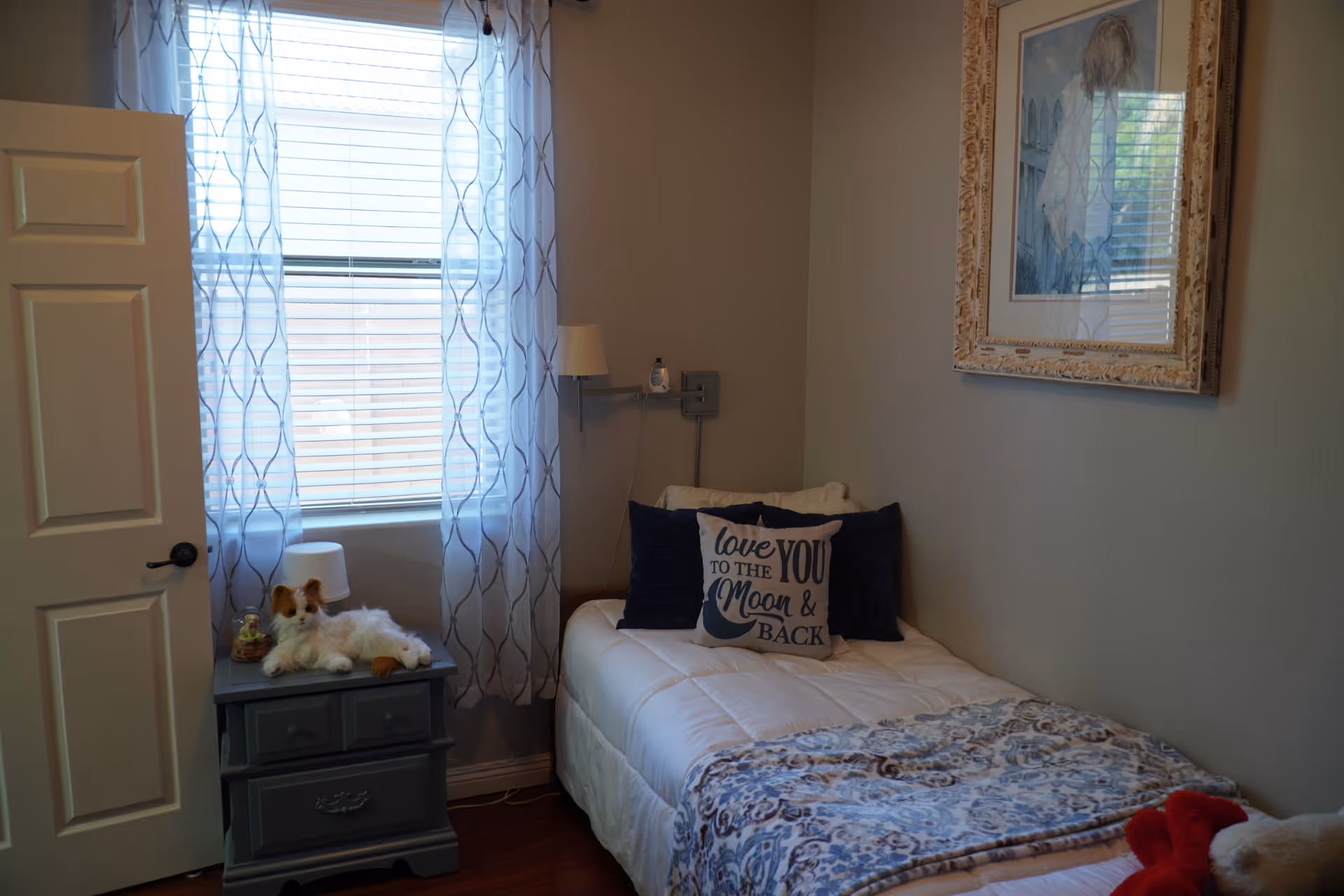 Small bedroom with a single bed, decorative pillows (one reading "love you to the moon & back"), a nightstand with a stuffed dog, and a window with patterned curtains.