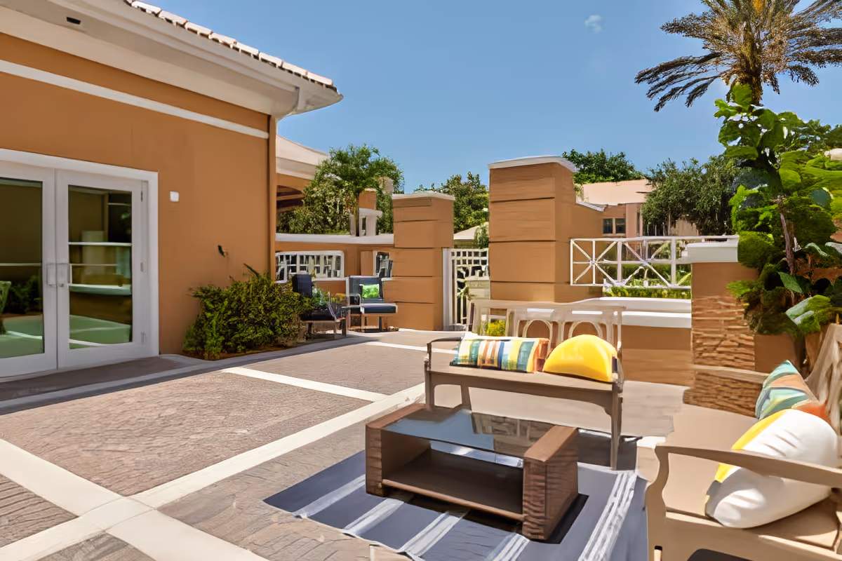 Outdoor patio area at Aston Gardens At Pelican Marsh featuring cushioned seating with colorful pillows, a glass-top coffee table on a striped rug, surrounded by plants and trees under a clear blue sky.