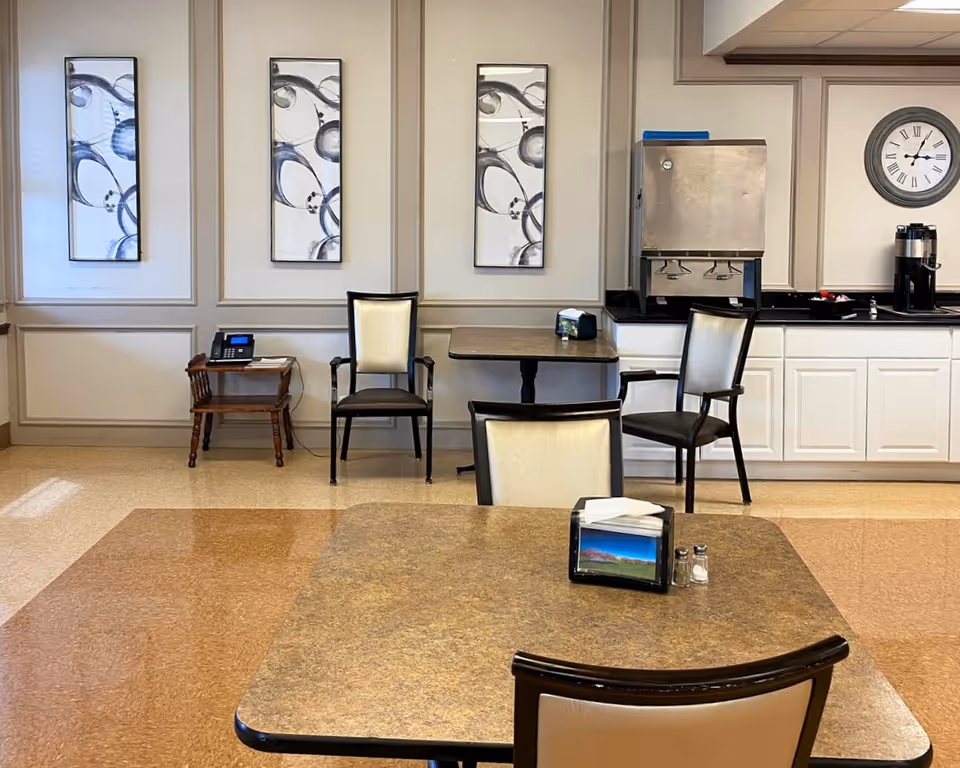 A dining area in a senior living facility with tables and chairs. There are three framed abstract art pieces on the wall, a small wooden side table with a telephone, a beverage dispenser, a coffee maker, and a wall clock showing the time as 3:10.