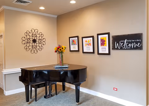 A black grand piano with a vase of sunflowers and a book on top is placed against a beige wall. The wall features decorative metal art, three framed colorful floral prints, and a sign that reads 'every family has a story Welcome to ours'. The room has recessed ceiling lights and a carpeted floor.