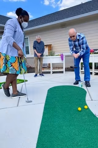 Three people playing miniature golf on an outdoor putting green at a senior living facility. One woman wearing a mask and colorful skirt is putting, while an elderly man and woman watch and wait their turn. The setting is outside near a building with beige siding and a raised garden bed.