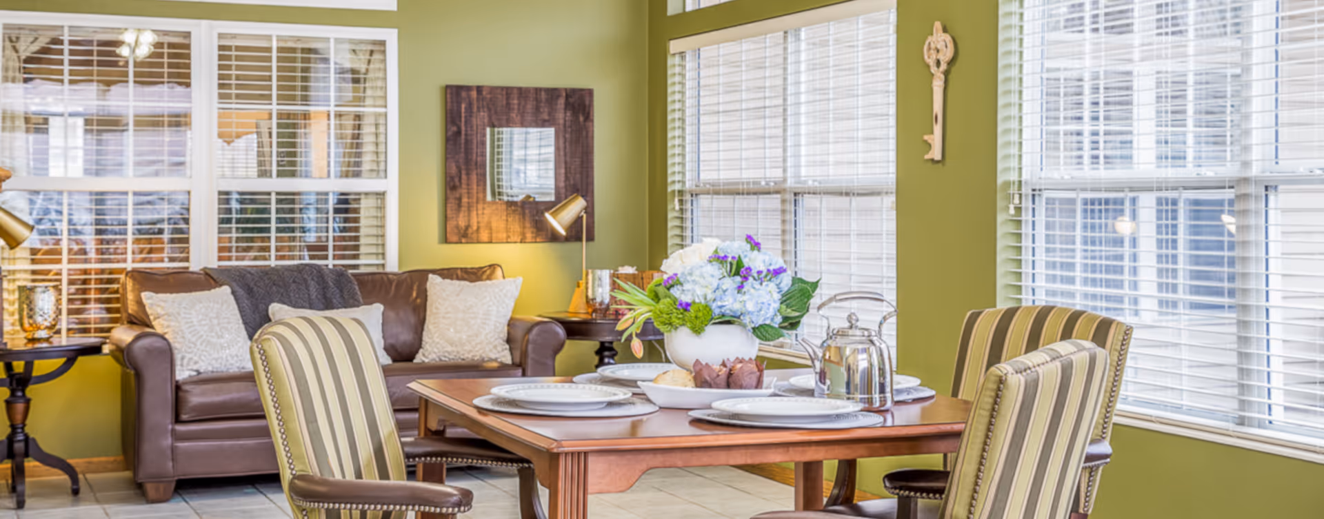 Sunlit dining area with a set wooden table and striped chairs next to a leather sofa in a green-walled living room.