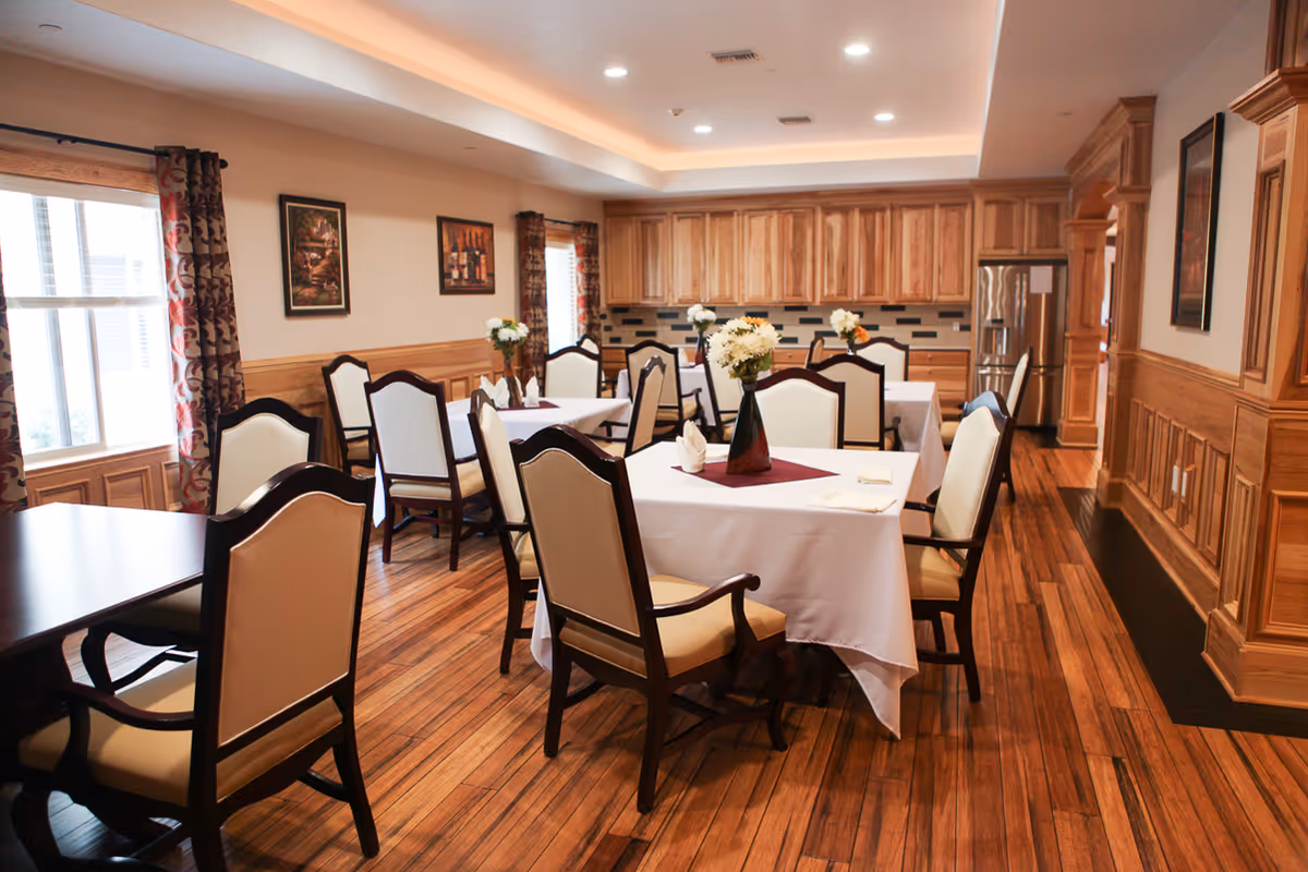 Dining room with several tables covered in white cloths, upholstered chairs, wood-paneled walls and a kitchen area with wooden cabinets.