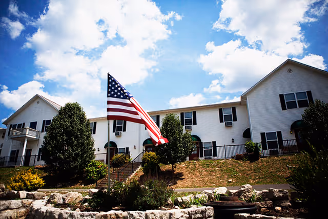 Exterior view of a two-story white building with multiple windows and green awnings, surrounded by trees and landscaping. An American flag is prominently displayed in the foreground against a partly cloudy blue sky.
