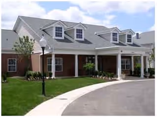 Exterior view of a senior living facility building with a covered entrance, brick and siding walls, multiple dormer windows on the roof, a curved driveway, a street lamp, and landscaped greenery.