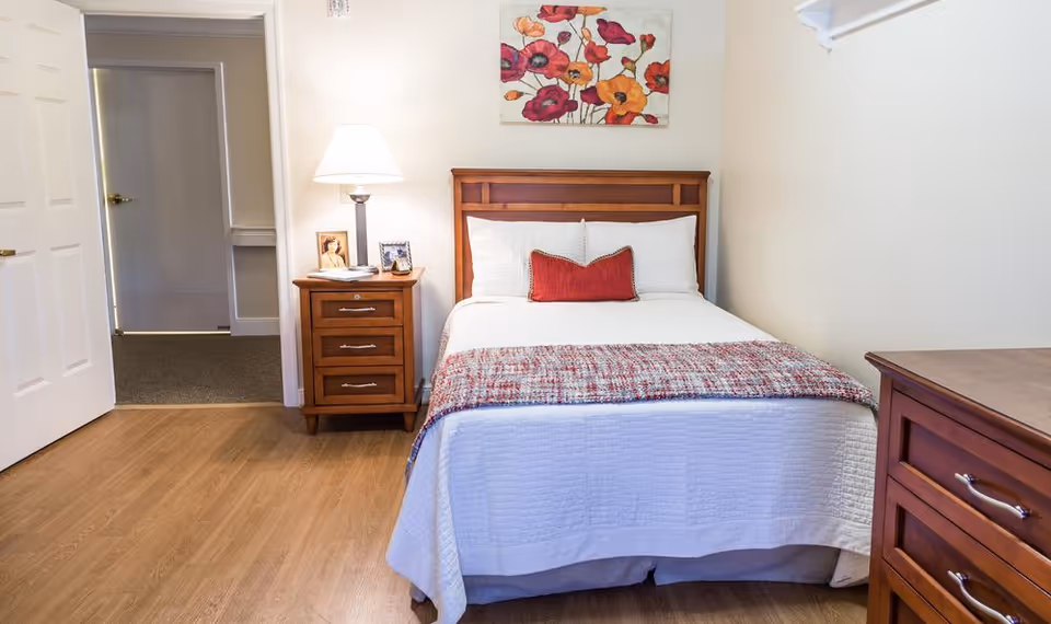 Cozy bedroom featuring a wooden bed frame with a white quilt and decorative red pillow, a bedside table with a lamp and personal items, and a floral wall art.