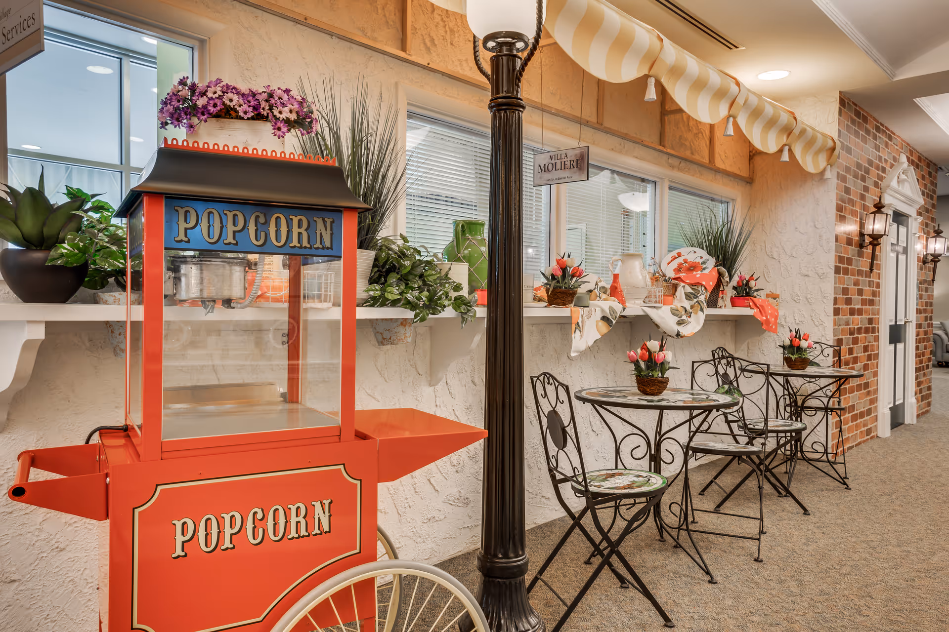 A red vintage popcorn cart sits next to wrought-iron bistro tables and decorative plants in a brightly decorated indoor corridor.