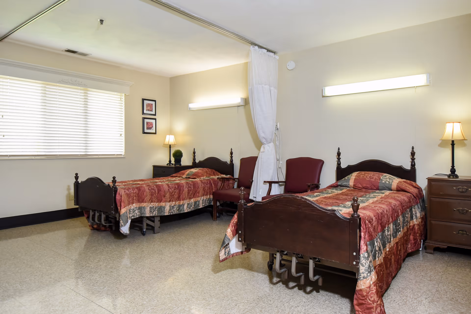 A room with two single hospital-style beds with wooden headboards and footboards, each covered with a red and green patterned quilt. Between the beds are two maroon chairs and a white privacy curtain. Each bed has a small wooden nightstand with a lamp. The room has a large window with closed blinds and two framed pictures on the wall.