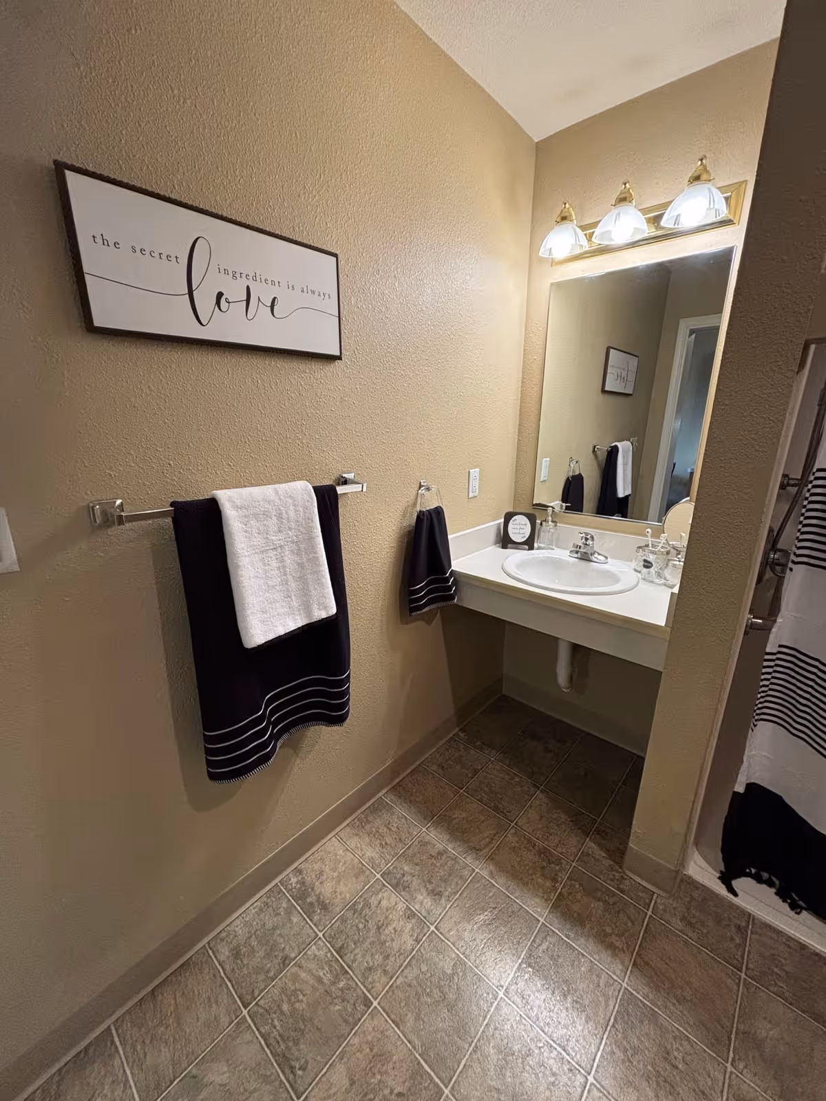 A bathroom with beige walls and tiled floor. There is a white sink with a large mirror above it and three light fixtures. Two towel racks hold black and white towels. A framed sign on the wall reads 'the secret ingredient is always love'. A shower curtain with black and white stripes is partially visible on the right.