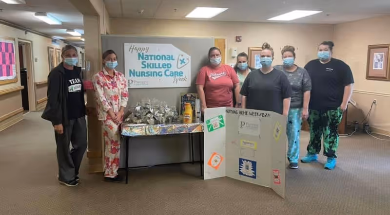 Seven healthcare workers wearing face masks stand in a hallway next to a table with gift bags and a sign that reads 'Happy National Skilled Nursing Care Week' at Prestige Care Center of Nebraska City. They are posing with a decorated poster board celebrating Nursing Home Week 2021.