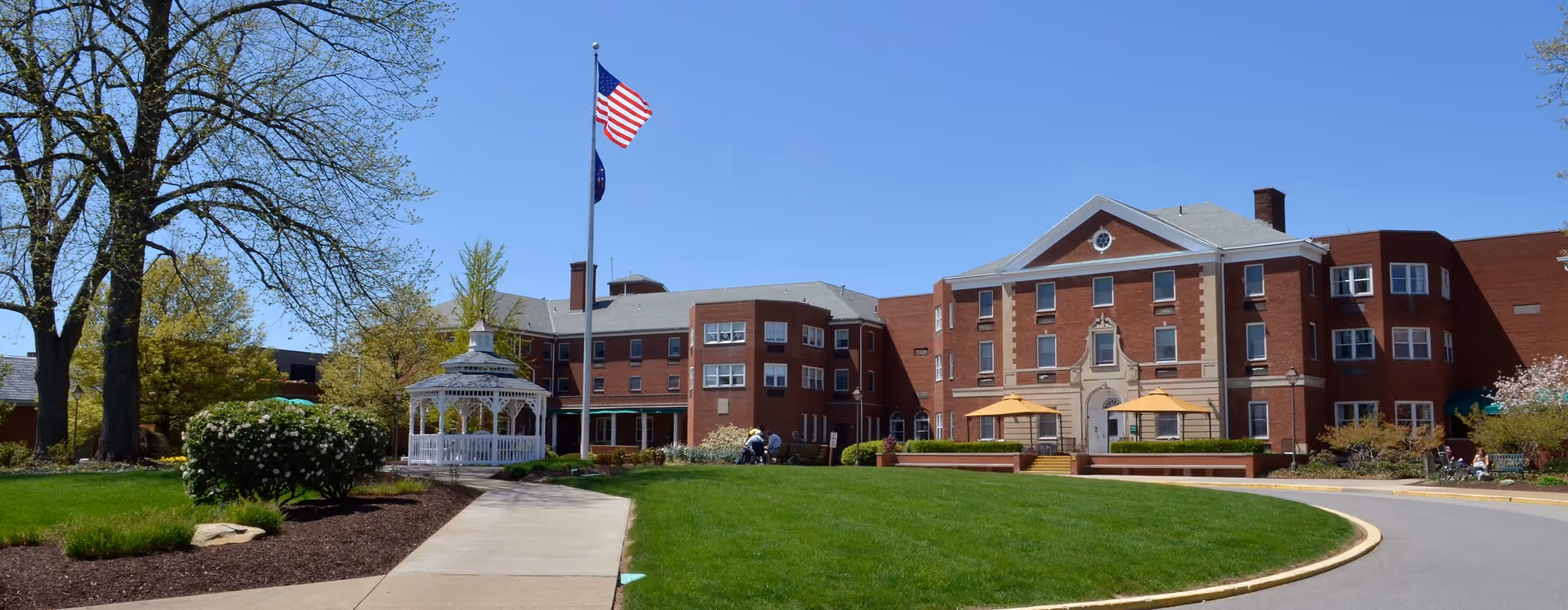 Exterior view of Asbury Heights senior living facility on a clear sunny day, featuring a large brick building with multiple windows, a white gazebo, a flagpole with the American flag, well-maintained green lawn, trees, and a curved driveway.