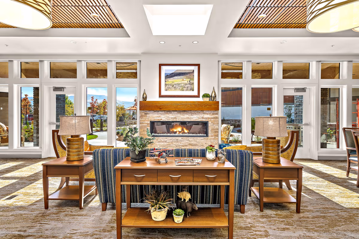 Bright communal lounge with upholstered chairs, side tables and a central fireplace in front of large windows.