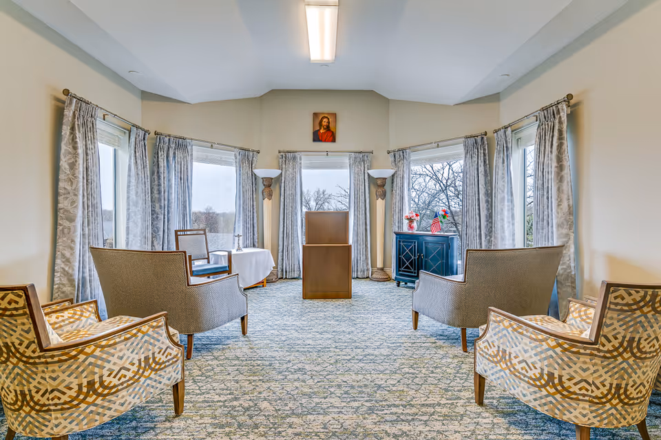 Bright common room with armchairs arranged around a central wooden lectern facing large windows and curtains.