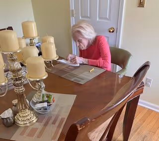 An elderly woman with white hair wearing a red long-sleeve shirt is seated at a wooden dining table, writing on a piece of paper. The table has placemats, a large decorative candle holder with multiple beige candles, a small bowl with packets, and a pencil. The room has light-colored walls and a closed white door in the background.