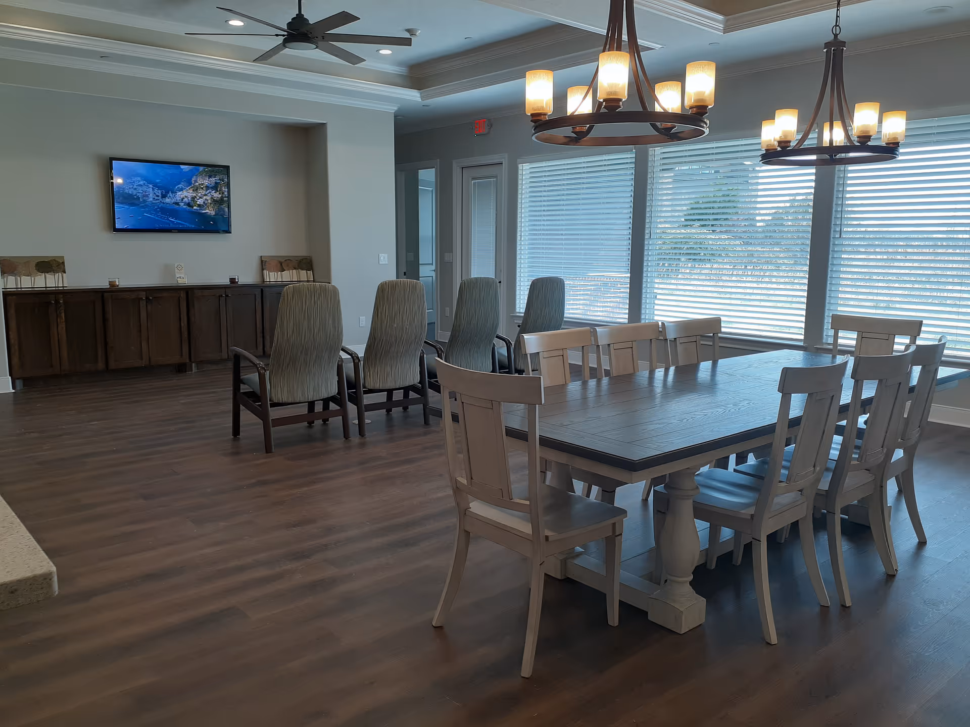 A spacious dining room with a large wooden dining table surrounded by eight chairs. Behind the dining area are four high-backed chairs facing a wall-mounted TV. The room has large windows with blinds, two ceiling light fixtures, and a ceiling fan. The floor is wooden, and there is a cabinet along the back wall.
