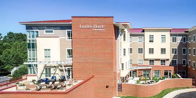 Exterior view of Linden House Assisted Living and Memory Care at Branchlands, showing a multi-story building with a brick and beige facade, outdoor seating area with patio furniture, and landscaped grounds under a clear sky.