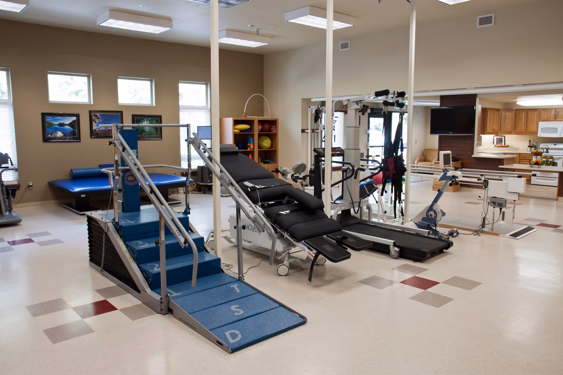 Physical therapy room with exercise equipment including steps, a treatment table, treadmill and therapy machines in a bright interior space.