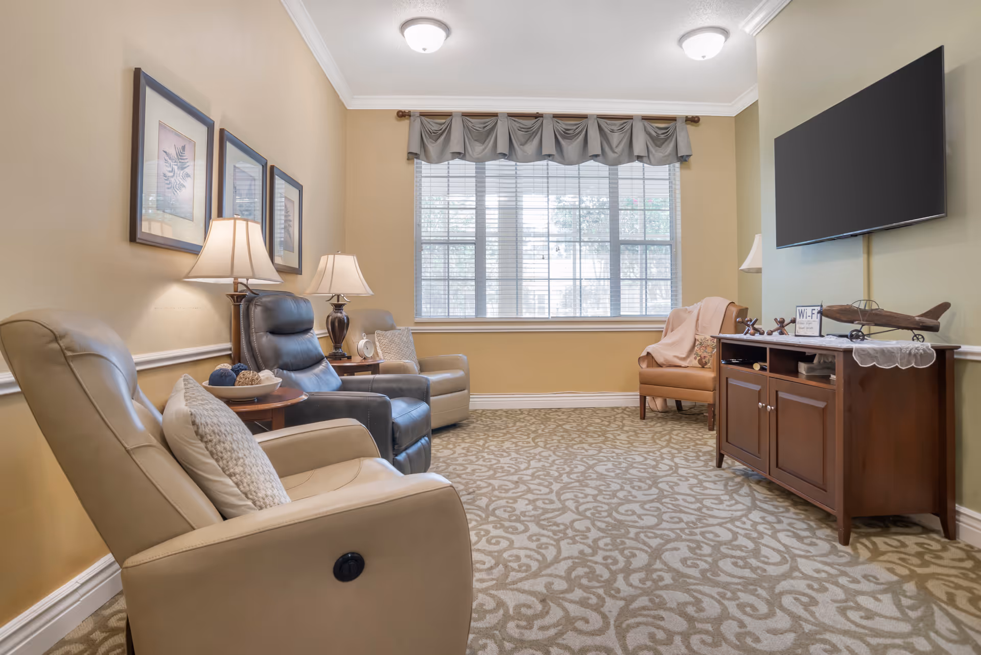 A cozy living room with three recliner chairs, two beige and one black, arranged around a wooden side table with lamps. There is a large window with blinds and a valance, a wall-mounted flat-screen TV above a wooden cabinet, and a single armchair with a blanket draped over it. The room has beige walls and patterned carpet flooring.