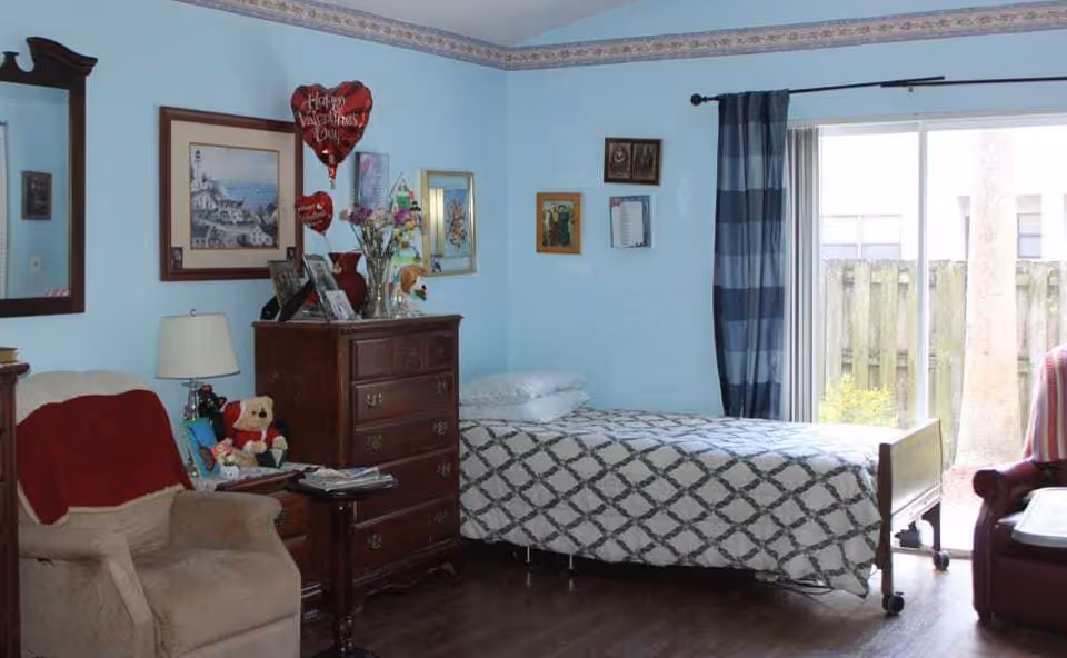 A cozy bedroom with light blue walls featuring a single bed with a patterned bedspread, a wooden chest of drawers adorned with framed photos and flowers, a beige armchair with a red throw, a small side table, and a large window with blue curtains letting in natural light.