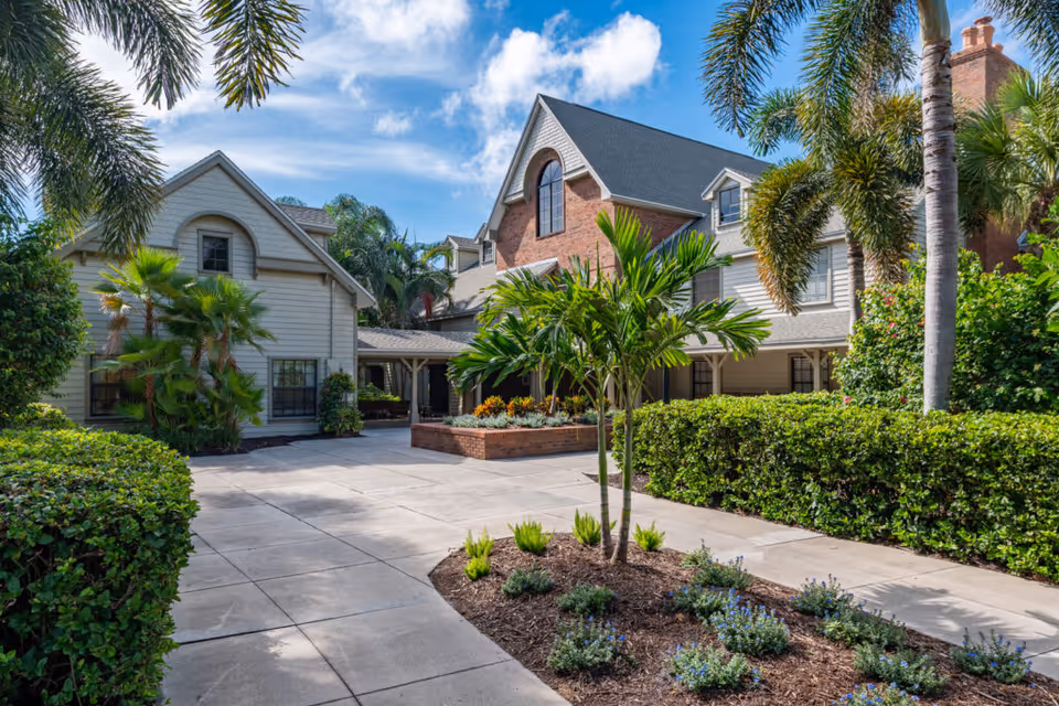 Exterior view of a senior living facility named South Port Square featuring a paved walkway, well-maintained landscaping with palm trees and shrubs, and a multi-story building with a mix of brick and siding under a partly cloudy blue sky.