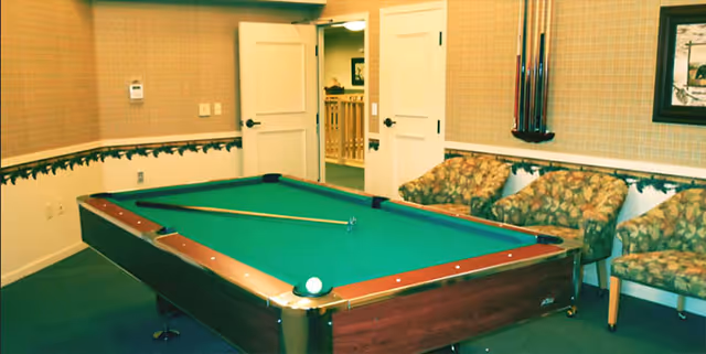 A recreation room with a pool table in the center, a cue stick resting on the table, three patterned armchairs along the wall, and a rack holding pool cues mounted on the wall.