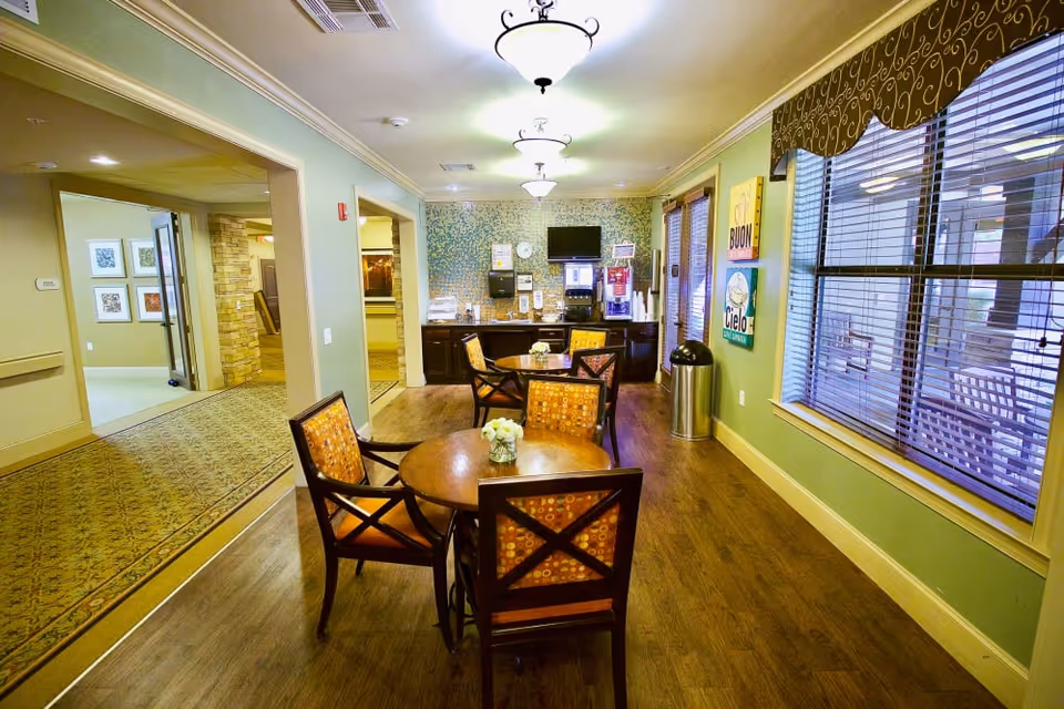 Bright common dining area with round wooden tables and cushioned chairs, a beverage station at the far wall, and large windows with blinds.