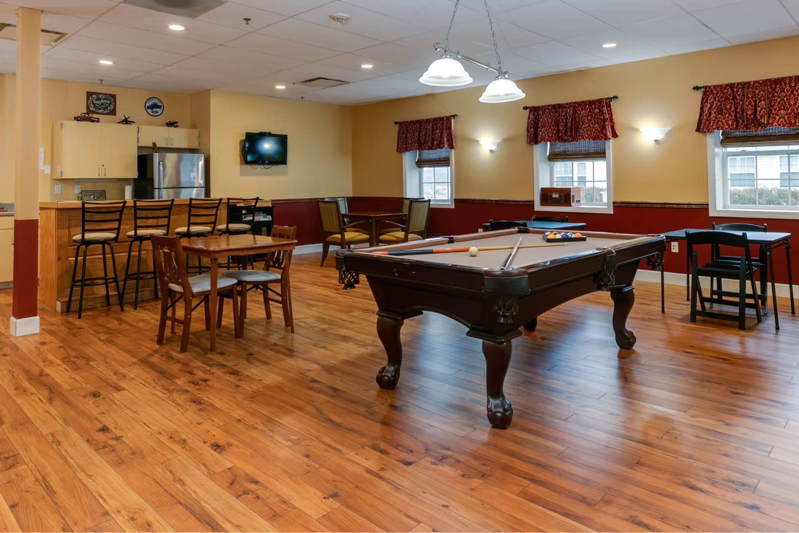 A recreational room with a pool table in the center, surrounded by several tables and chairs. There is a small kitchen area with a refrigerator and bar stools on the left side. The room has wooden flooring, yellow and red walls, three windows with red curtains, and ceiling lights.