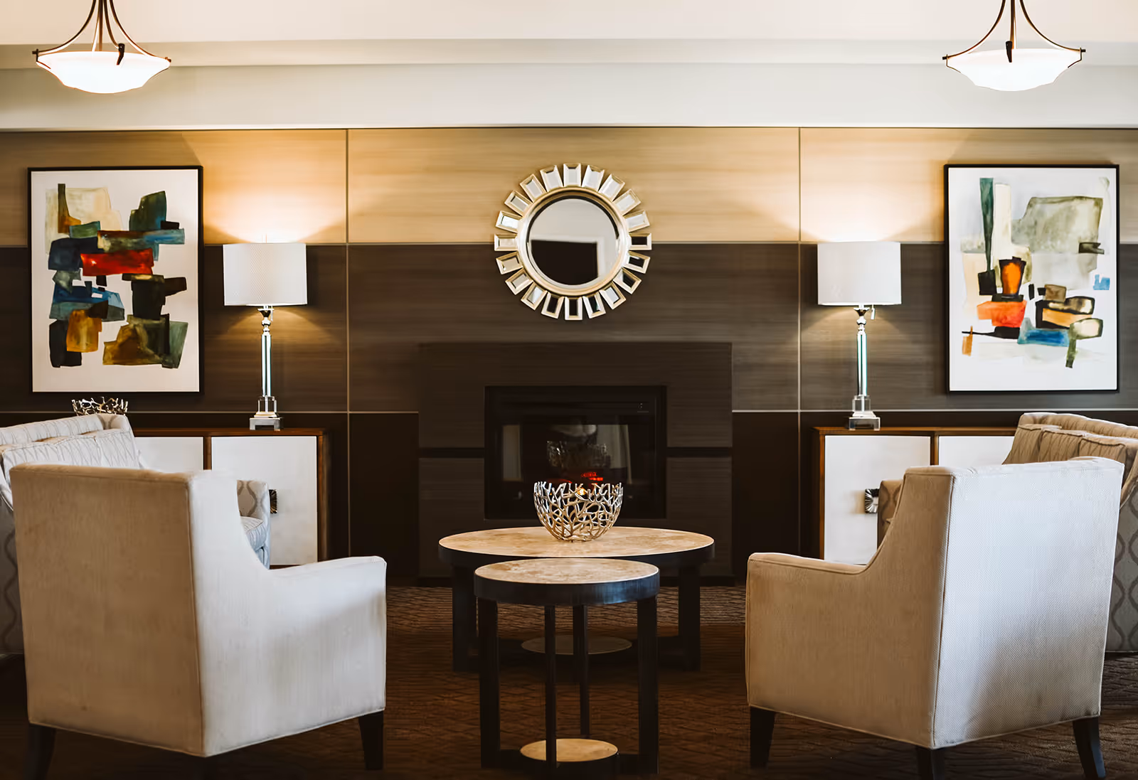 A cozy living room area with two beige armchairs facing each other across two round nested coffee tables. Behind the tables is a modern fireplace with a decorative round mirror above it. On either side of the fireplace are side tables with lamps and abstract art paintings hanging on the wall.