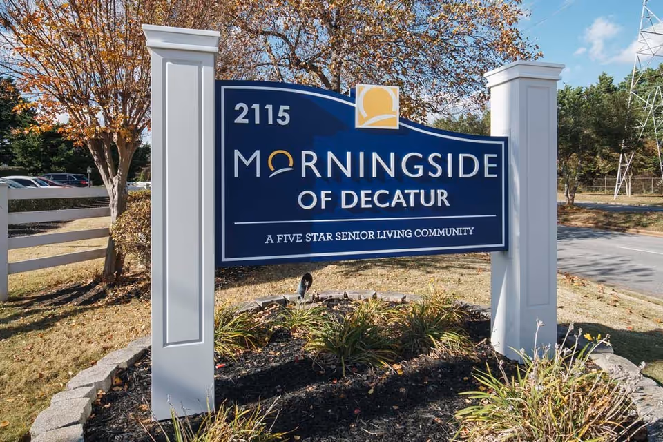 Outdoor sign for Morningside of Decatur, a five star senior living community, with the address 2115. The sign is blue with white and yellow lettering, mounted between two white posts, surrounded by landscaping and trees with autumn leaves.