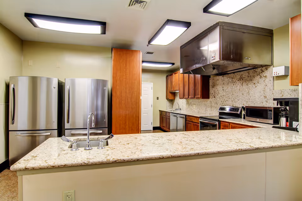 Modern kitchen with two stainless steel refrigerators, a large granite countertop with a built-in sink and faucet, wooden cabinets, a stainless steel stove with an overhead vent, a microwave, and a coffee maker.