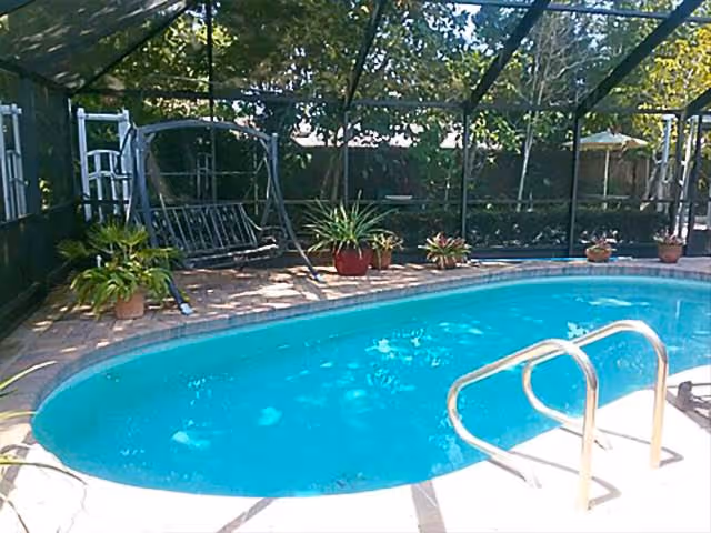 Outdoor swimming pool with clear blue water surrounded by a screened enclosure. There are several potted plants around the pool area and a metal swing bench on the left side.